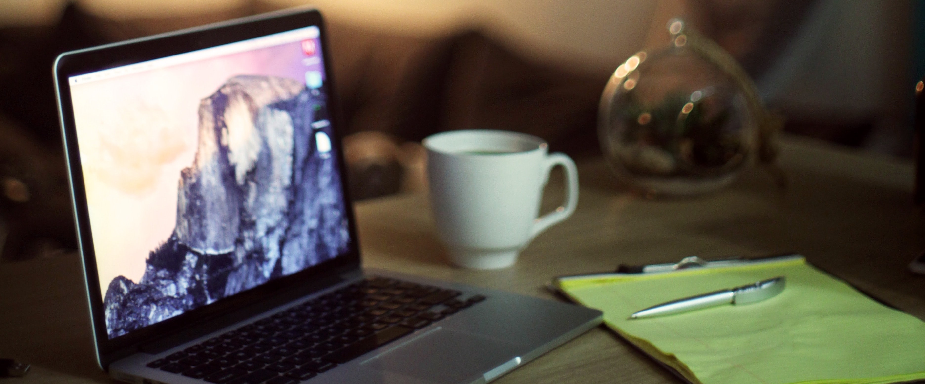 A laptop, mug, pen and paper sit on a desk